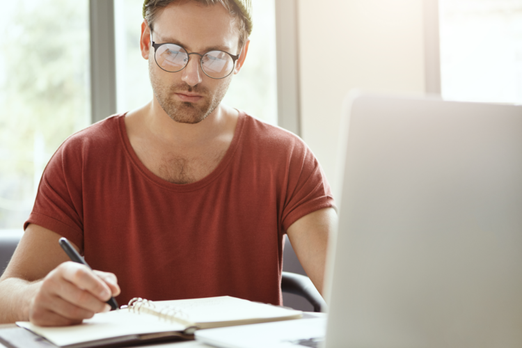 A man sat at a desk writing in a notebook in front of a laptop