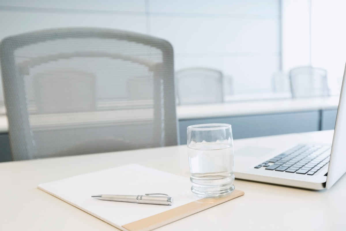 A desk with a glass, a pen, a laptop, and a paper pad