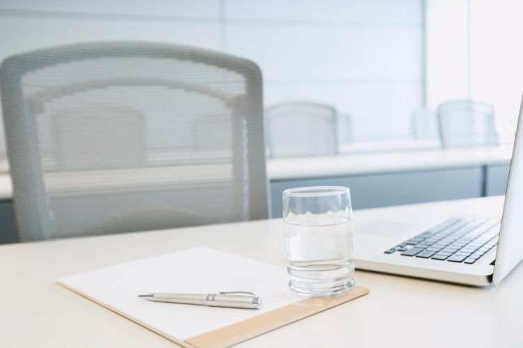 A desk with a glass, a pen, a laptop, and a paper pad