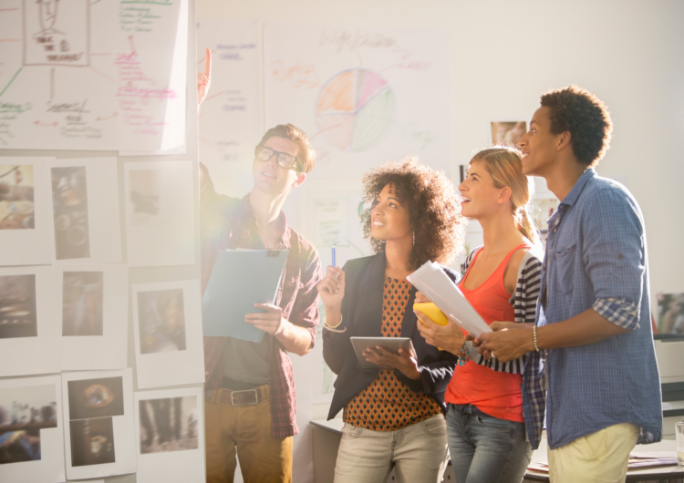 A group of casually dressed people in an office setting looking at a wall of notes