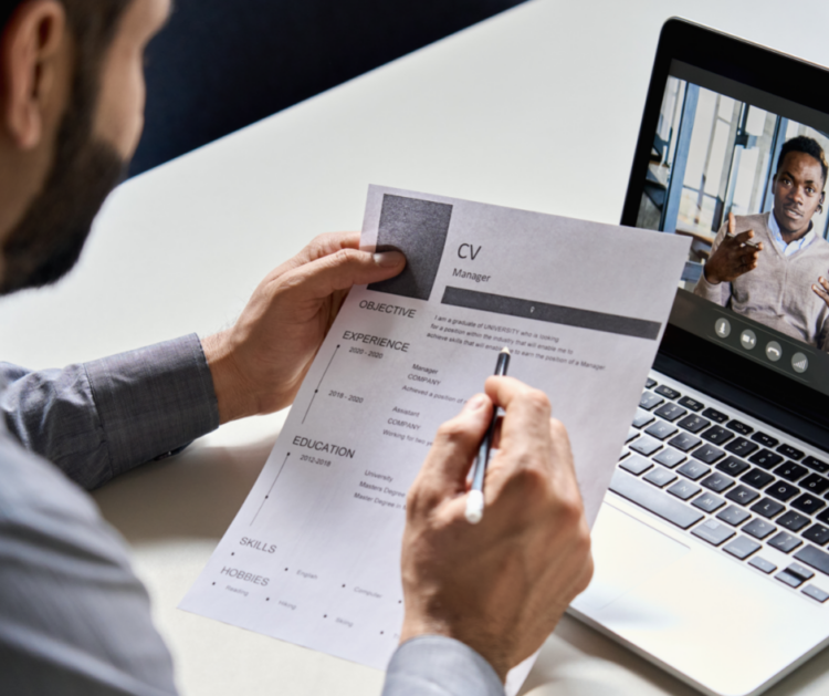 A man reading a CV with another man sat on a zoom call on a laptop in front of him