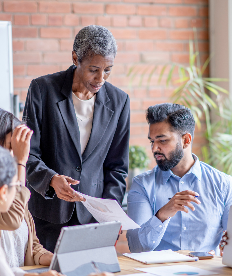 A older lady in an office with work colleagues showing them a spreadsheet.