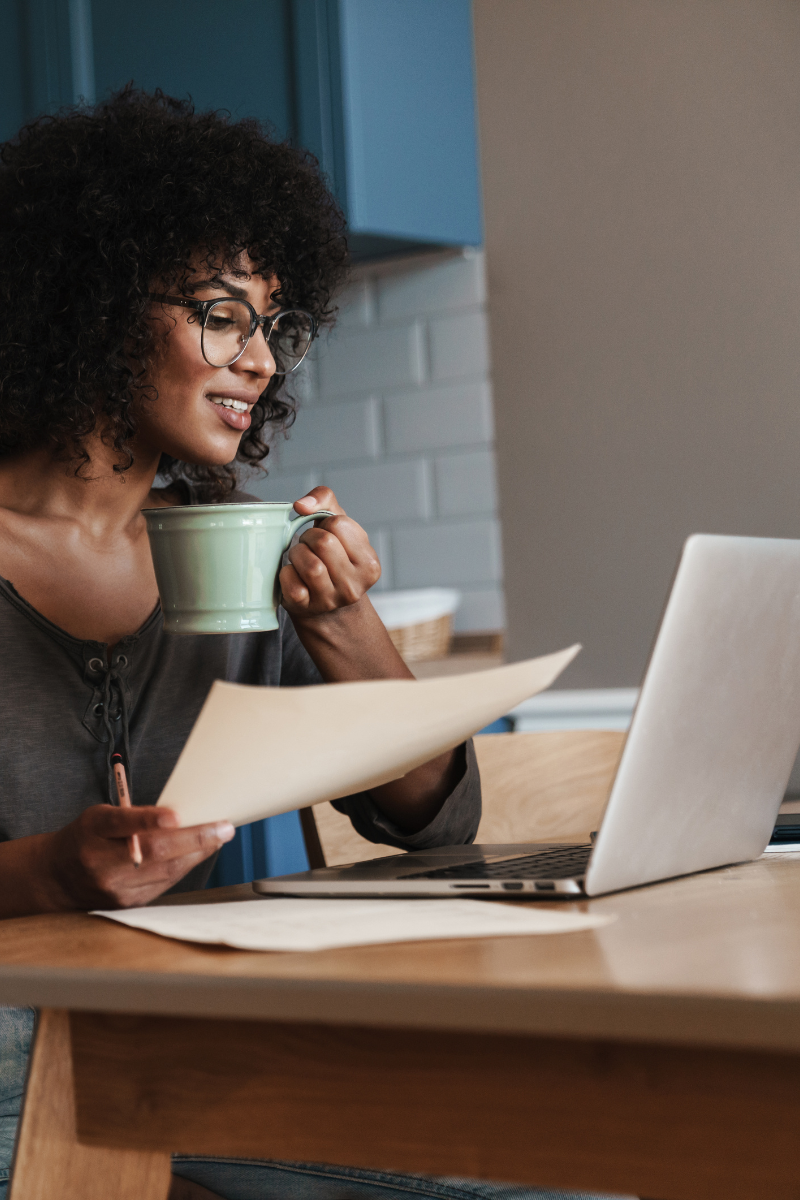 A work from Home woman drinking tea with some paper in hand whilst smiling at her laptop.